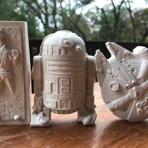 K&ouml;nnte beinhalten: Drei cremefarbene Star Wars-Figuren: Han Solo in Karbonit, R2-D2 und der Millennium Falke. Die Figuren bestehen aus einem hellen Material und werden auf einer Holzoberfl&auml;che pr&auml;sentiert.