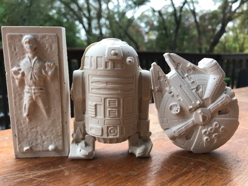 K&ouml;nnte beinhalten: Drei cremefarbene Star Wars-Figuren: Han Solo in Karbonit, R2-D2 und der Millennium Falke. Die Figuren bestehen aus einem hellen Material und werden auf einer Holzoberfl&auml;che pr&auml;sentiert.