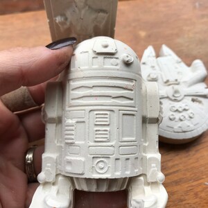 K&ouml;nnte beinhalten: Eine wei&szlig;e R2-D2-Figur wird in einer Hand gehalten, mit einem Han Solo in Karbonit-Relief und einem Millennium Falcon-Modell im Hintergrund. Die Figuren sind alle wei&szlig; und scheinen aus Gips oder einem &auml;hnlichen Material zu bestehen.