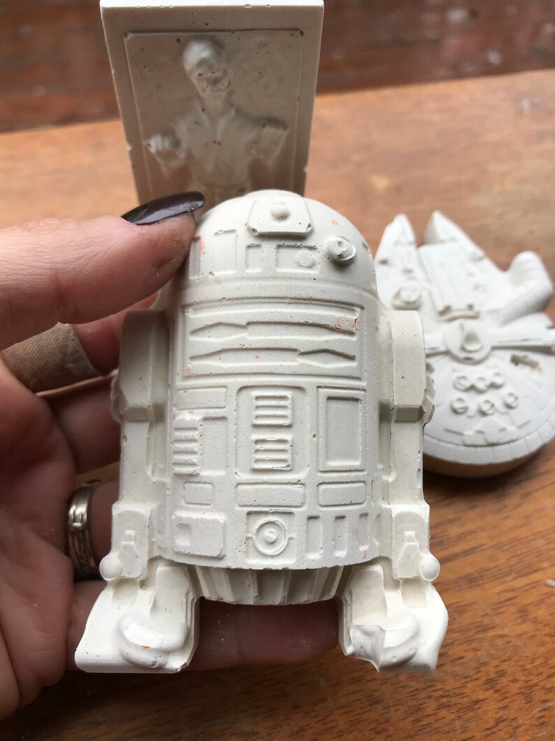 K&ouml;nnte beinhalten: Eine wei&szlig;e R2-D2-Figur wird in einer Hand gehalten, mit einem Han Solo in Karbonit-Relief und einem Millennium Falcon-Modell im Hintergrund. Die Figuren sind alle wei&szlig; und scheinen aus Gips oder einem &auml;hnlichen Material zu bestehen.