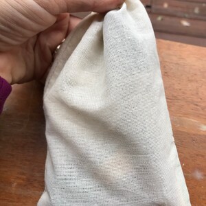 K&ouml;nnte beinhalten: Ein kleiner, cremefarbener Beutel aus Musselin mit Kordelzug, gehalten von einer Hand mit dunklem Nagellack. Der Beutel ist gef&uuml;llt und leicht abgerundet, was darauf hindeutet, dass er kleine Gegenst&auml;nde enth&auml;lt. Der Hintergrund ist eine Holzoberfl&auml;che.