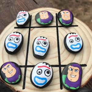 May include: A hand-painted tic-tac-toe game on a wooden slice. The game pieces are painted rocks featuring characters from the film Toy Story. The game board has black lines, and the rocks are painted with the faces of Buzz Lightyear and Forky.