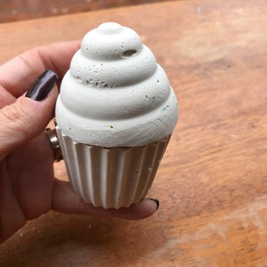 May include: A light gray cupcake-shaped object with a textured, swirled top and a ribbed base. The object is held in a hand against a wooden background. The cupcake appears to be made of concrete or a similar material.