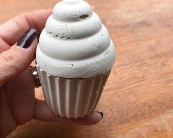 Puede incluir: Un objeto en forma de cupcake de color gris claro con una parte superior texturizada y arremolinada y una base acanalada. El objeto se sostiene en una mano sobre un fondo de madera. El cupcake parece estar hecho de hormigón o un material similar.