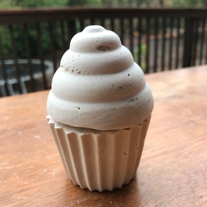 May include: A light gray cupcake-shaped container with a textured surface. The top resembles frosting, and the base has a fluted design. The container is made of a solid material and sits on a wooden surface.