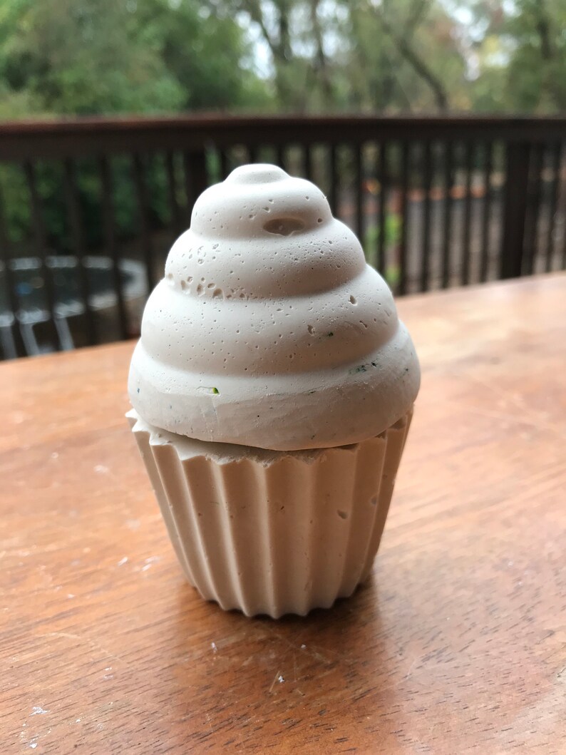 May include: A light gray cupcake-shaped container with a textured surface. The top resembles frosting, and the base has a fluted design. The container is made of a solid material and sits on a wooden surface.
