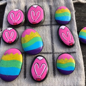 Könnte beinhalten: Ein handgemachtes Tic-Tac-Toe-Spielset mit bemalten Steinen. Die Spielsteine ​​umfassen Steine, die mit rosa Hasengesichtern und Regenbogen-Eier-Designs bemalt sind. Das Spiel befindet sich auf einem grauen Leinenbeutel.