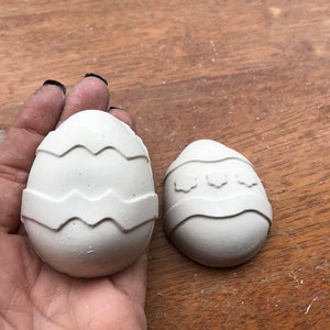 May include: Two white, egg-shaped decorations with textured, zig-zag and floral designs. The eggs are displayed on a wooden surface. One egg is larger than the other.