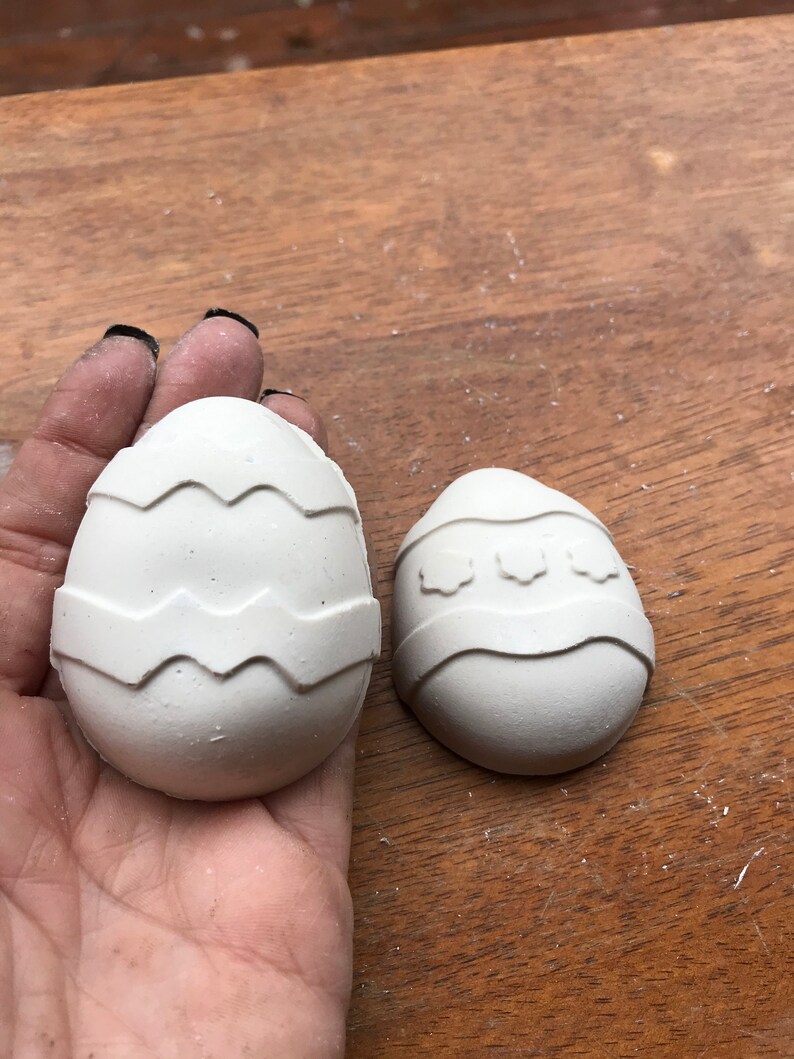 May include: Two white, egg-shaped decorations with textured, zig-zag and floral designs. The eggs are displayed on a wooden surface. One egg is larger than the other.