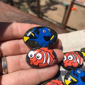 May include: Hand-painted river rocks featuring characters from the animated film "Finding Nemo." The rocks are painted with bright colors, including orange, white, blue, and yellow. The characters include Nemo and Dory.