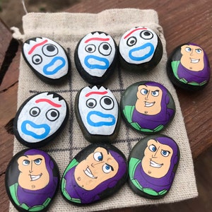 May include: Hand-painted stone tic-tac-toe game featuring characters from the Toy Story franchise. The game includes nine stones, with five stones painted as Buzz Lightyear and four as Forky. The game pieces are on a small, beige cloth bag.