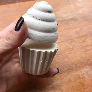 May include: A white, textured cupcake-shaped object with a fluted base and a swirled top. The cupcake is held in a hand against a wooden surface. The object appears to be made of a porous material.
