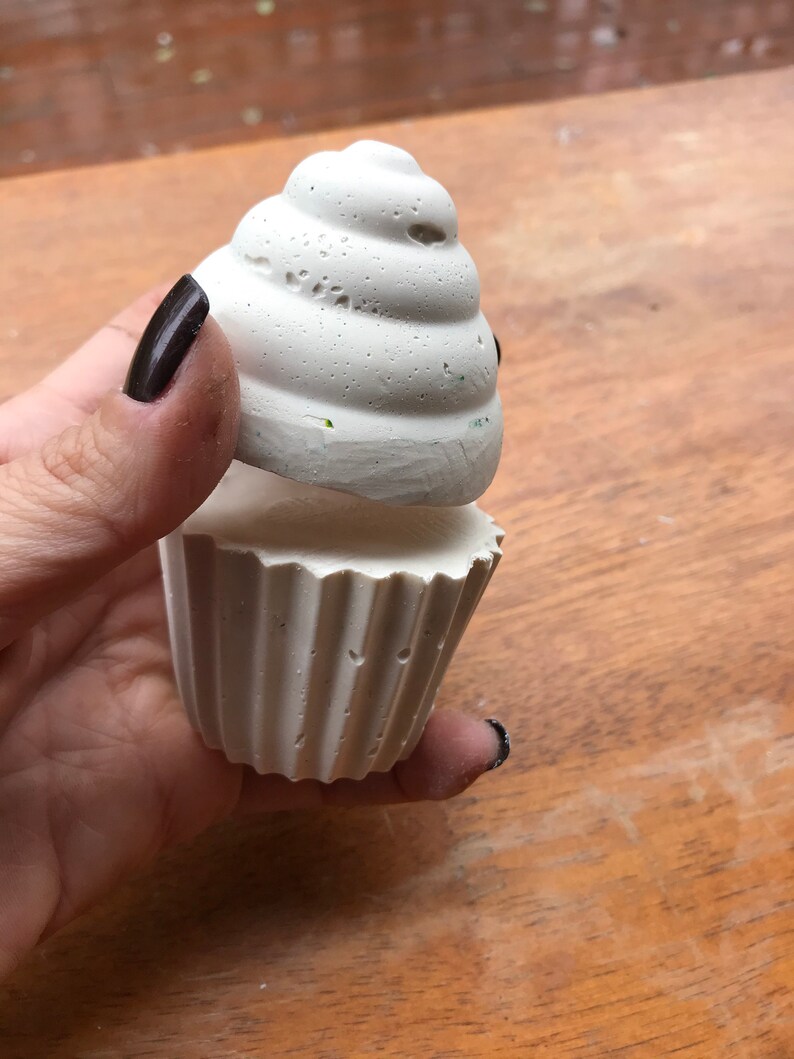 May include: A white, textured cupcake-shaped object with a fluted base and a swirled top. The cupcake is held in a hand against a wooden surface. The object appears to be made of a porous material.