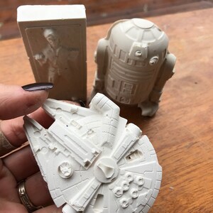 K&ouml;nnte beinhalten: Hellwei&szlig;e Star Wars Figuren. Enth&auml;lt einen Millennium Falken, R2-D2 und eine Figur in Karbonit. Der Millennium Falke hat detaillierte Verkleidungen und ein zentrales Cockpit. R2-D2 ist ein runder Droide mit rechteckigen Paneelen.