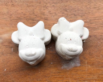 Puede incluir: Dos figuras blancas con forma de Minnie Mouse con lazos. Las figuras están hechas de un material similar al yeso y descansan sobre una superficie de madera. Las figuras de Minnie Mouse tienen una cara sonriente.