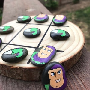 May include: A tic-tac-toe game set on a wooden board. The game pieces are painted rocks featuring cartoon characters in green, purple, and black. The board is a circular wooden slice with black lines.