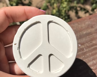 Puede incluir: Un emblema redondo blanco roto con el símbolo de la paz. El símbolo de la paz está en relieve y centrado en la superficie circular. El emblema se sostiene en una mano con esmalte de uñas rosa, con un fondo borroso verde y marrón.