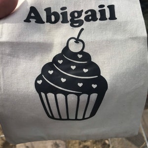 May include: A white tote bag with the name "Abigail" printed in black, above a black cupcake design. The cupcake has heart-shaped decorations and a cherry on top. The bag is made of a light-colored fabric.