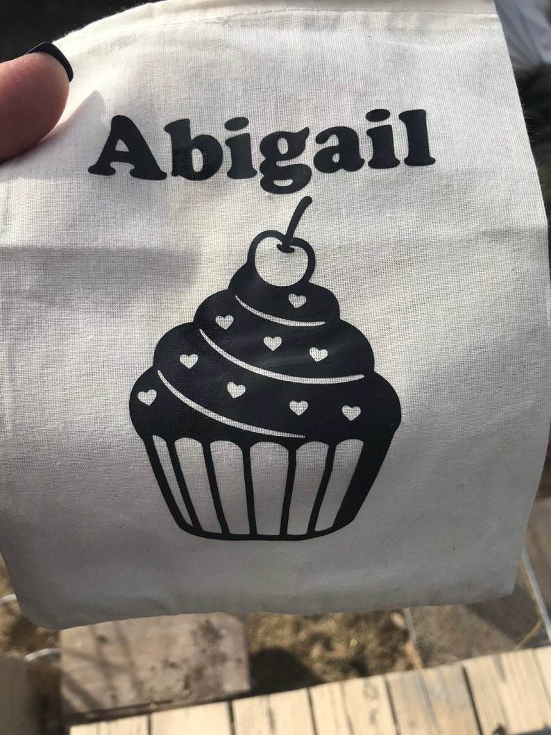 May include: A white tote bag with the name "Abigail" printed in black, above a black cupcake design. The cupcake has heart-shaped decorations and a cherry on top. The bag is made of a light-colored fabric.