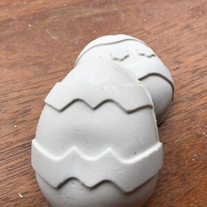 May include: Two off-white, egg-shaped decorations with a zig-zag pattern around the middle. The eggs are resting on a wooden surface. The decorations are likely for Easter.