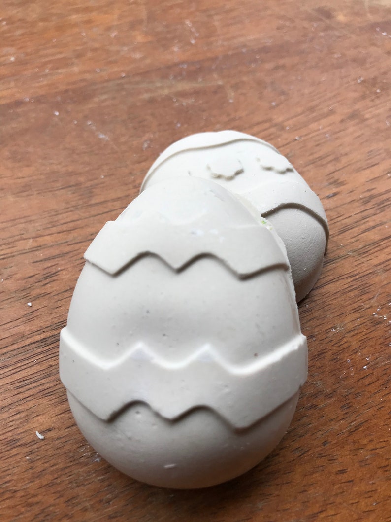 May include: Two off-white, egg-shaped decorations with a zig-zag pattern around the middle. The eggs are resting on a wooden surface. The decorations are likely for Easter.