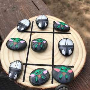Puede incluir: Juego de tres en raya sobre una rodaja de madera. Las piezas son piedras pintadas con el casco mandaloriano y Grogu, también conocido como Baby Yoda, personajes de la franquicia Star Wars. El tablero tiene líneas negras.