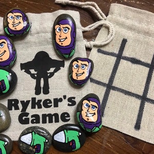 May include: A handcrafted game set with painted rocks depicting Buzz Lightyear and alien characters. The set includes a drawstring bag printed with "Ryker's Game" and a separate bag with a tic-tac-toe grid. The rocks are painted in bright colours.