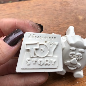 May include: A white, rectangular Toy Story stamp with the Disney Pixar logo. The stamp is held in a hand with dark nail polish. A white Mr. Potato Head figure is visible to the right.