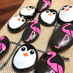 Puede incluir: Piedras decorativas pintadas a mano con diseños de pingüinos y flamencos. Los pingüinos tienen vientres blancos, cuerpos negros y picos naranjas. Los flamencos son rosas con detalles negros. Las piedras están dispuestas sobre una tela beige.