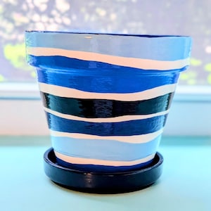 Puede incluir: Una maceta cónica con acabado brillante que presenta rayas horizontales en tonos azul claro, azul medio, azul oscuro, negro y blanco. La maceta se asienta sobre un platillo circular negro a juego, creando un conjunto visualmente coherente.