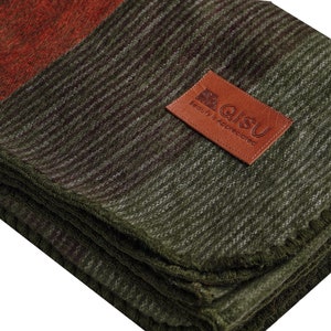 May include: Folded blanket with a color block design in shades of green, brown, and rust. A brown leather patch with the text "QISU Beauty is Appreciated" is sewn on the blanket. The blanket has a textured, woven appearance.