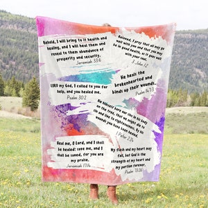 May include: A colorful throw blanket with inspirational bible verses printed on it. The blanket is made of soft, fuzzy fabric and features a watercolor-like design with shades of pink, purple, blue, and green. The verses are printed in black text and include quotes about healing, hope, and faith.