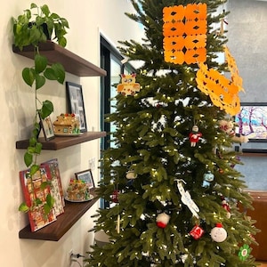 May include: A decorated Christmas tree with orange paper ornaments, a small owl ornament, and white lights. The tree is in front of a wall with wooden shelves holding plants, pictures, and small gingerbread houses.