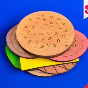 Puede incluir: Un juego de posavasos de madera con forma de hamburguesa con un pan marrón, carne rosa, queso amarillo, lechuga verde y un pan marrón en la parte inferior. El pan está decorado con pequeñas semillas de sésamo amarillas.