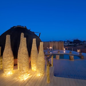 May include: A wooden deck with a swimming pool and a thatched roof hut in the background. There are five tall, cylindrical light fixtures made of recycled materials, illuminating the space.