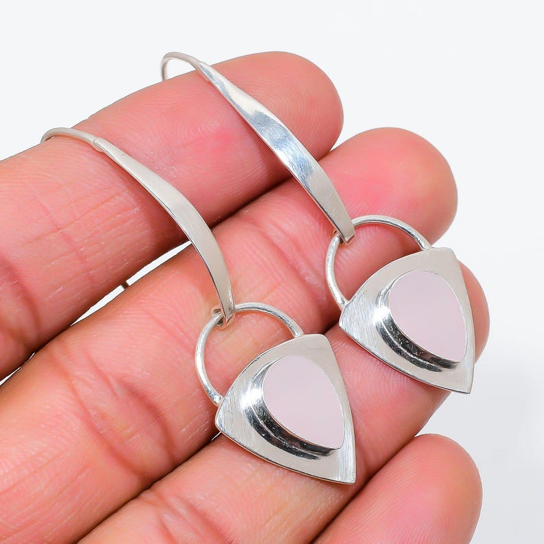 K&ouml;nnte beinhalten: Ein Paar silberne Ohrh&auml;nger mit einem gebogenen Metalldraht-Haken, der an einem dreieckigen Silberrahmen befestigt ist. Jeder Rahmen enth&auml;lt einen glatten, blassrosa, tropfenf&ouml;rmigen Stein in der Mitte, was ein ausgewogenes geometrisches Design ergibt.