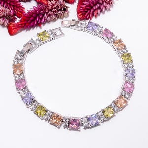 Könnte beinhalten: Ein silberfarbenes Metallarmband mit einer Reihe quadratisch geschliffener, mehrfarbiger Edelsteine in Gelb-, Rosa-, Violett- und Pfirsichtönen. Jeder große quadratische Stein wird von kleinen, klaren, runden Akzentsteinen in einem Kettendesign flankiert.