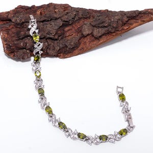 Könnte beinhalten: Ein silberfarbenes Armband mit einer Reihe oval geschliffener olivgrüner Steine, die sich mit kleinen klaren weißen Kristallen abwechseln. Die Metallglieder sind in einem geschwungenen Muster angeordnet und verfügen über einen Kastenverschluss.