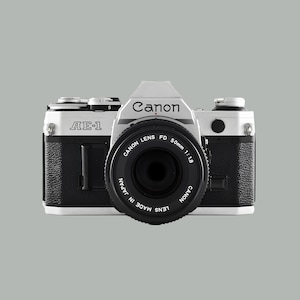 May include: A Canon AE-1 camera with a 50mm lens. The camera body is black and white, with the Canon logo and AE-1 model name visible. The lens is labelled "CANON LENS FD 50mm 1:1.8" and "MADE IN JAPAN".