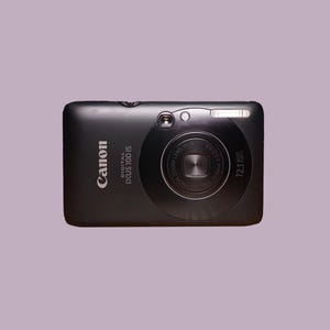 May include: Black Canon Digital IXUS 100 IS digital camera. The camera has a zoom lens and a flash. The camera has the text "Canon" and "DIGITAL IXUS 100 IS" printed on the front.