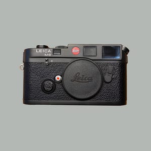 Puede incluir: Una cámara telemétrica Leica M6 negra con un cuerpo texturizado y una tapa de objetivo. La cámara tiene el logotipo de Leica en rojo y blanco, y el nombre del modelo es visible. La cámara está sobre un fondo gris.