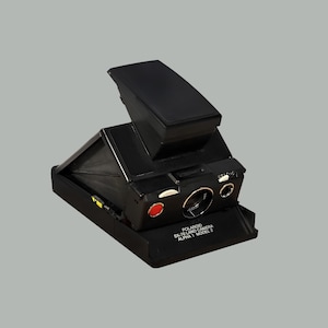 May include: A black Polaroid SX-70 Land Camera Alpha 1 Model 2, with a pop-up viewfinder and a red button. The camera is on a gray background. The camera is a vintage instant film camera.
