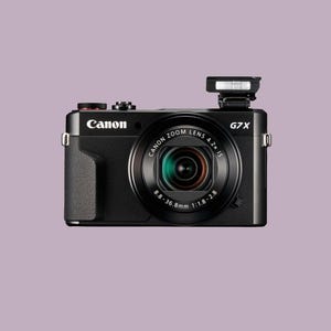 May include: A black Canon G7X digital camera with a zoom lens and a pop-up flash. The camera has a silver lens ring and a red button on top. The Canon logo is visible on the front.
