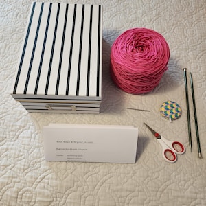 May include: A beginner knitting kit with 3 projects. The kit includes a box, pink yarn, knitting needles, a yarn needle, a measuring tape, and scissors. The box has black and white stripes. The card says "Knit Knack & Beyond presents... Beginner Knit Kit with 3 Projects."