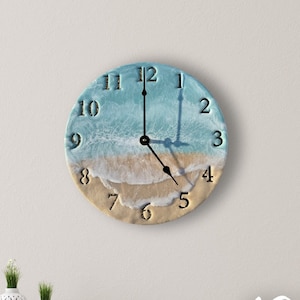 May include: A 10 inch round clock with a beach scene design. The clock face features a blue ocean with white waves and a sandy beach. The numbers are black and the hands are black.