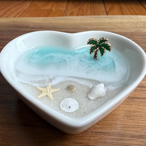 May include: A white heart-shaped trinket dish with a beach scene. The dish features a blue and white resin ocean, a starfish, seashells, and a palm tree charm. The dish is on a wooden surface.