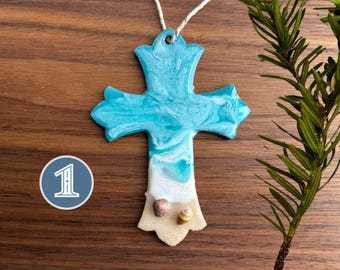 Op de afbeelding: Een blauwe en witte kruisversiering met een strandscene en schelpen. Het kruis is gemaakt van hars en heeft een wit touwtje om op te hangen.