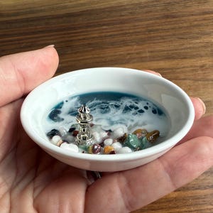 Op de afbeelding: Een kleine, witte keramische schaal met een miniatuur vuurtorenscène. De scène omvat een zilveren vuurtoren, blauwe en witte hars die de oceaan voorstelt en kleine kiezelstenen. De schaal wordt in een hand gehouden.