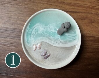 May include: A round white ceramic tray featuring a resin art design that mimics a beach scene. The surface displays a sandy beige area with two small seashells and a turquoise blue ocean section with white foam waves and a small grey seal figure.