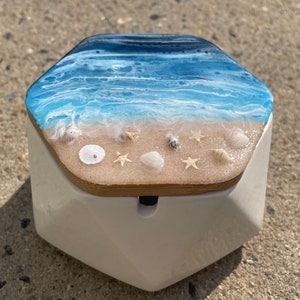 May include: A white hexagonal ceramic box with a lid featuring a resin ocean scene. The lid has a blue and white ocean with white foam waves and a sandy beach with seashells and starfish.